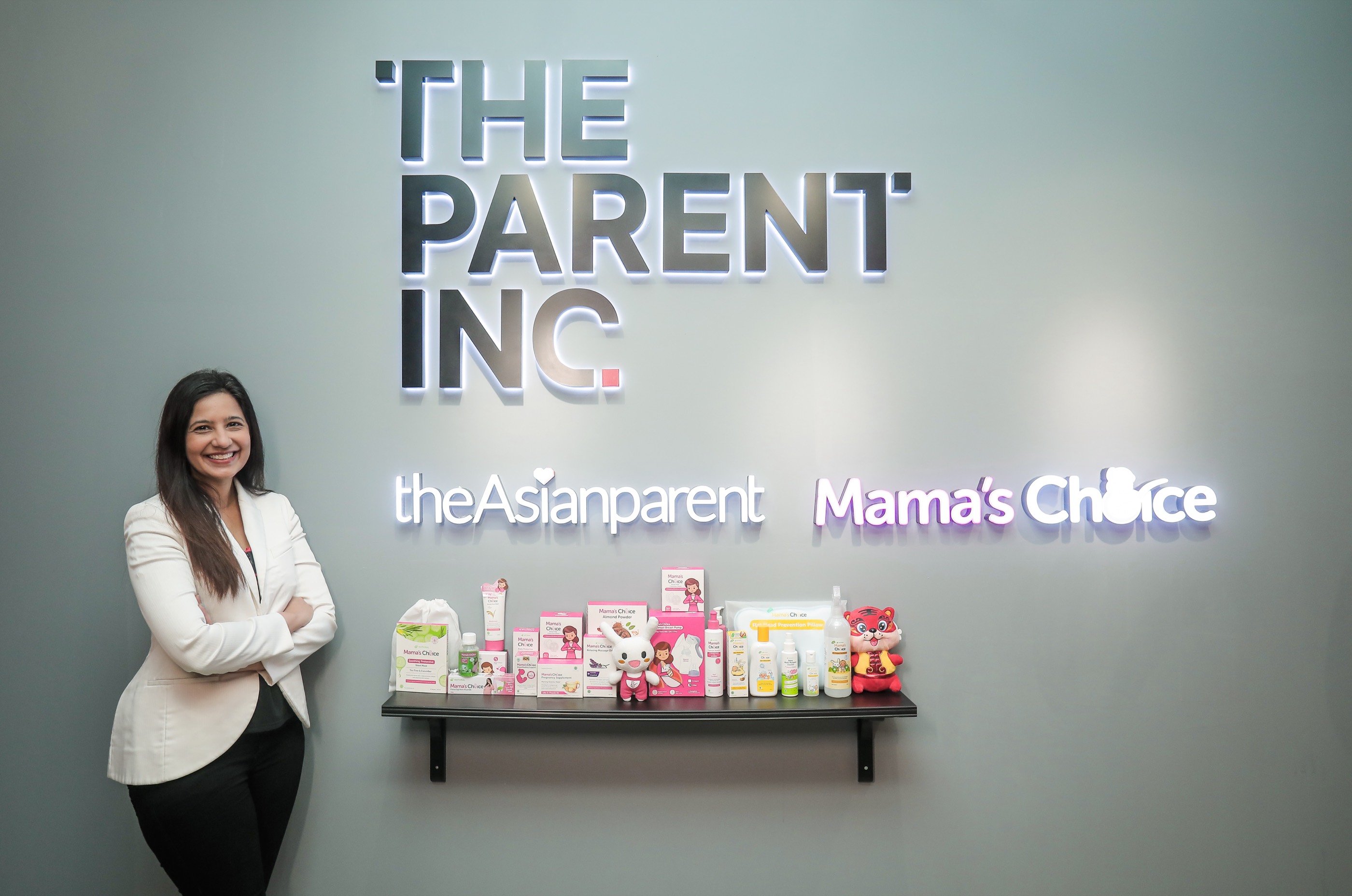 Roshni Mahtani Cheung beside The Parent Inc sign, with theAsianparent and Mama's Choice logos and a shelf of Mama's Choice products